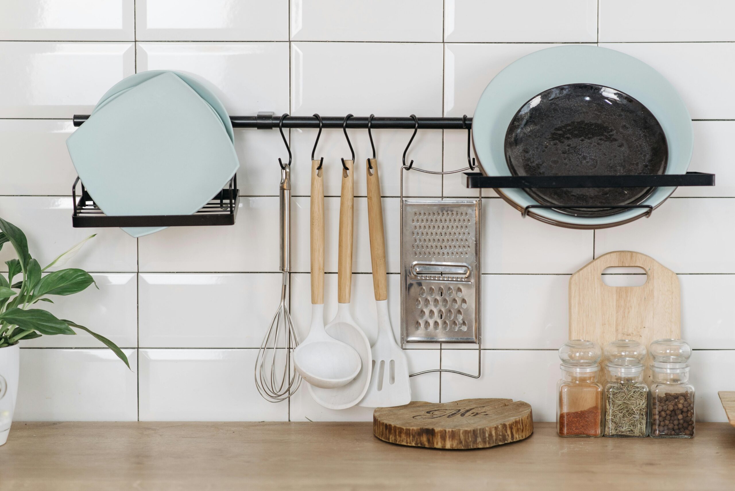 A photo of a kitchen counter. Mounted on the tile backsplash is a rack holding plates and various kitchen utensils. On the counter is a wood trivet and an assortment of jarred spices.