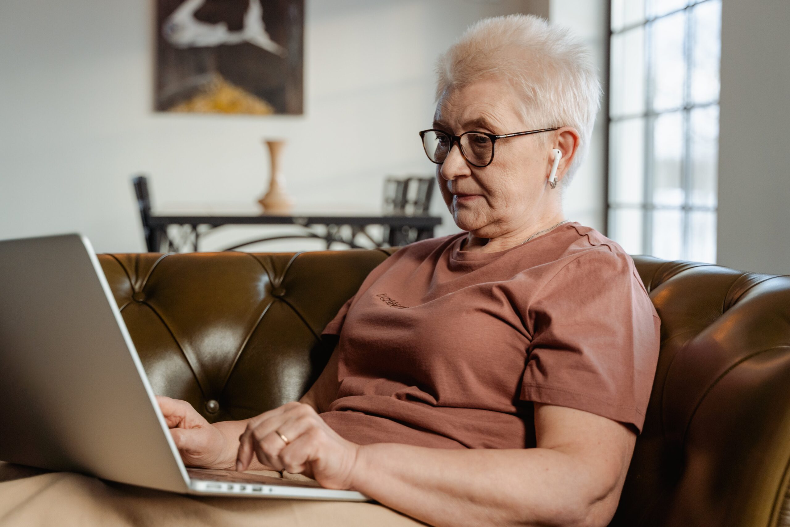 An older women lays on a couch looking at a laptop that is perched on her legs. She is wearing wireless earbuds and glasses, and her hands are poised over the keyboard as if she is about to type.