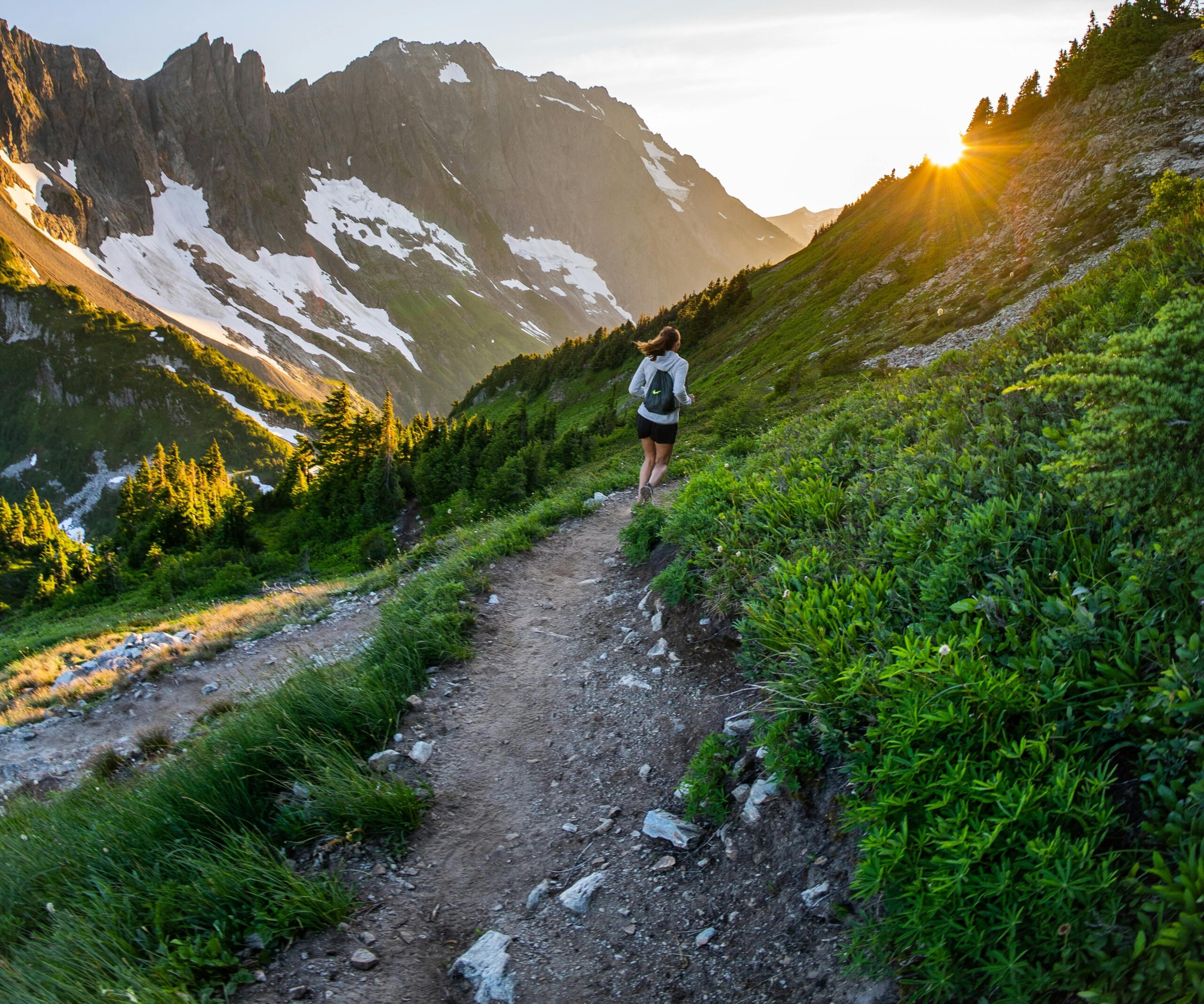 A photograph of a lone person runs on a trail with a spectacular backdrop of craggy mountains. The trail is on the side of the steep hillside, and the sun is slipping out of view.
