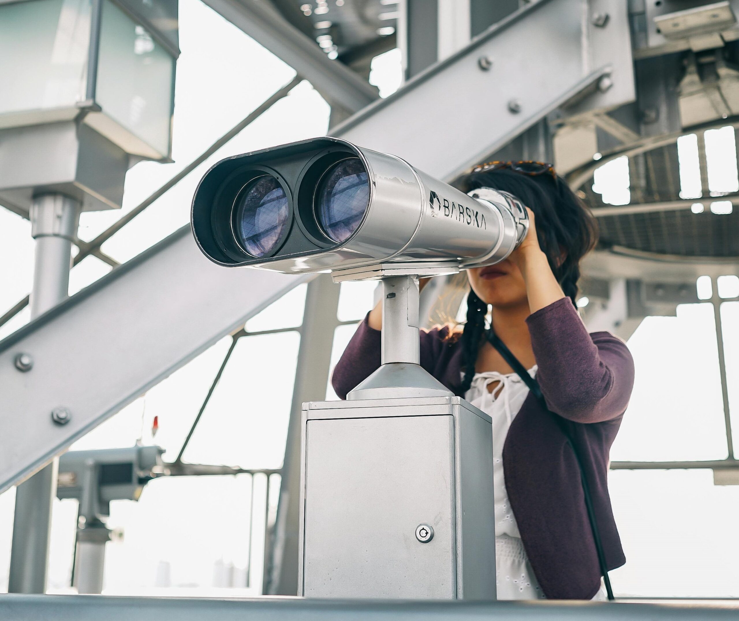 A photograph of a person wearing a purple cardigan and a white blouse looks out from a large binocular stand on the viewing deck of a tall building. The binoculars are pointed towards the left side of the photograph.