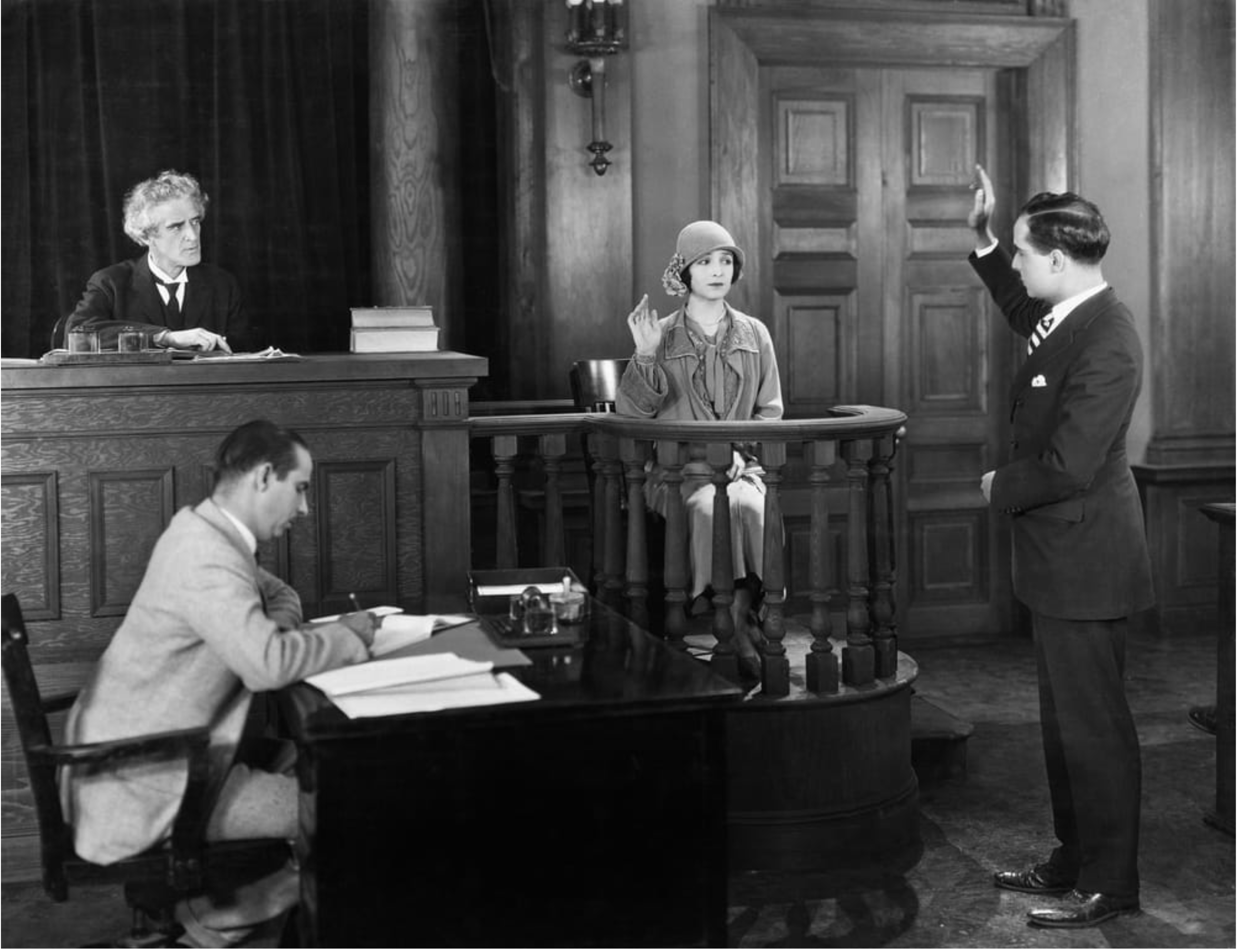 Black and white photo of an old criminal trial where a woman is being sworn in as a witness by the lawyer as the judge looks on and the court reporter writes what is being said.